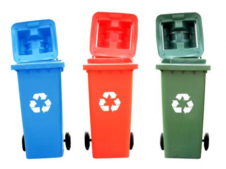The image displays three wheeled rubbish bins positioned in a row on a paved driveway or outdoor surface, each with an open hinged lid. The first bin on the left is bright blue with a smooth, matte finish, featuring a white recycling symbol on the front; the middle bin is red, also with a matte surface, displaying a similar white recycling icon, while the third bin on the right is green with a matching matte finish and the same recycling logo. All three bins are made of plastic, with visible handles near the lid edges and black wheels at the base, positioned in a well-lit outdoor environment. The background is plain and neutral, with no specific environmental or urban features visible, emphasizing the clear and orderly presentation of the waste containers. This arrangement reflects typical rubbish and recycling bin setups seen in residential or commercial waste management contexts, similar to those handled by services such as Commercial Waste Removal Plaistow. The image highlights the different bin colors used for separate waste streams, consistent with local waste disposal practices in the Plaistow area of London, inferred from the webpage context and postcode.
