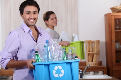 Two individuals are standing in a domestic kitchen setting, with the focus on a young man in the foreground holding a blue recycling bin filled with various clear glass bottles, some with blue and green tinting, and a few plastic bottles, all arranged upright inside the bin which features a white recycling symbol on the front. The man is wearing a light purple shirt with rolled-up sleeves and has a friendly smile. In the background, a woman with dark hair tied back, dressed in a light-colored top, is slightly blurred and appears to be organizing or packing items into a green plastic box on a kitchen countertop. The kitchen has wooden cabinets, a tiled backsplash, and neutral-toned walls, suggesting a typical residential environment. The scene is well-lit, with natural or artificial lighting highlighting the textures of the glass bottles and the wooden furniture, emphasizing the context of waste sorting or rubbish collection, which aligns with services offered by Commercial Waste Removal Plaistow. The image reflects a focus on recycling activities within a household, subtly associated with rubbish removal services in the Plaistow area.